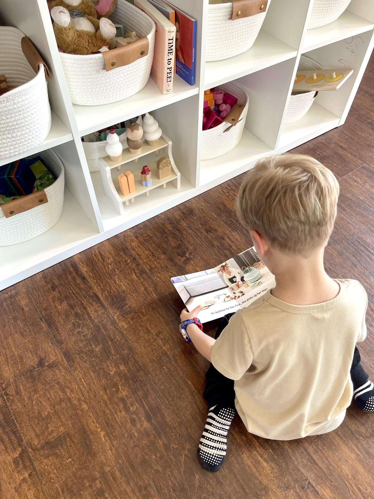 A child sitting on the floor reading a picture book beside low open shelves